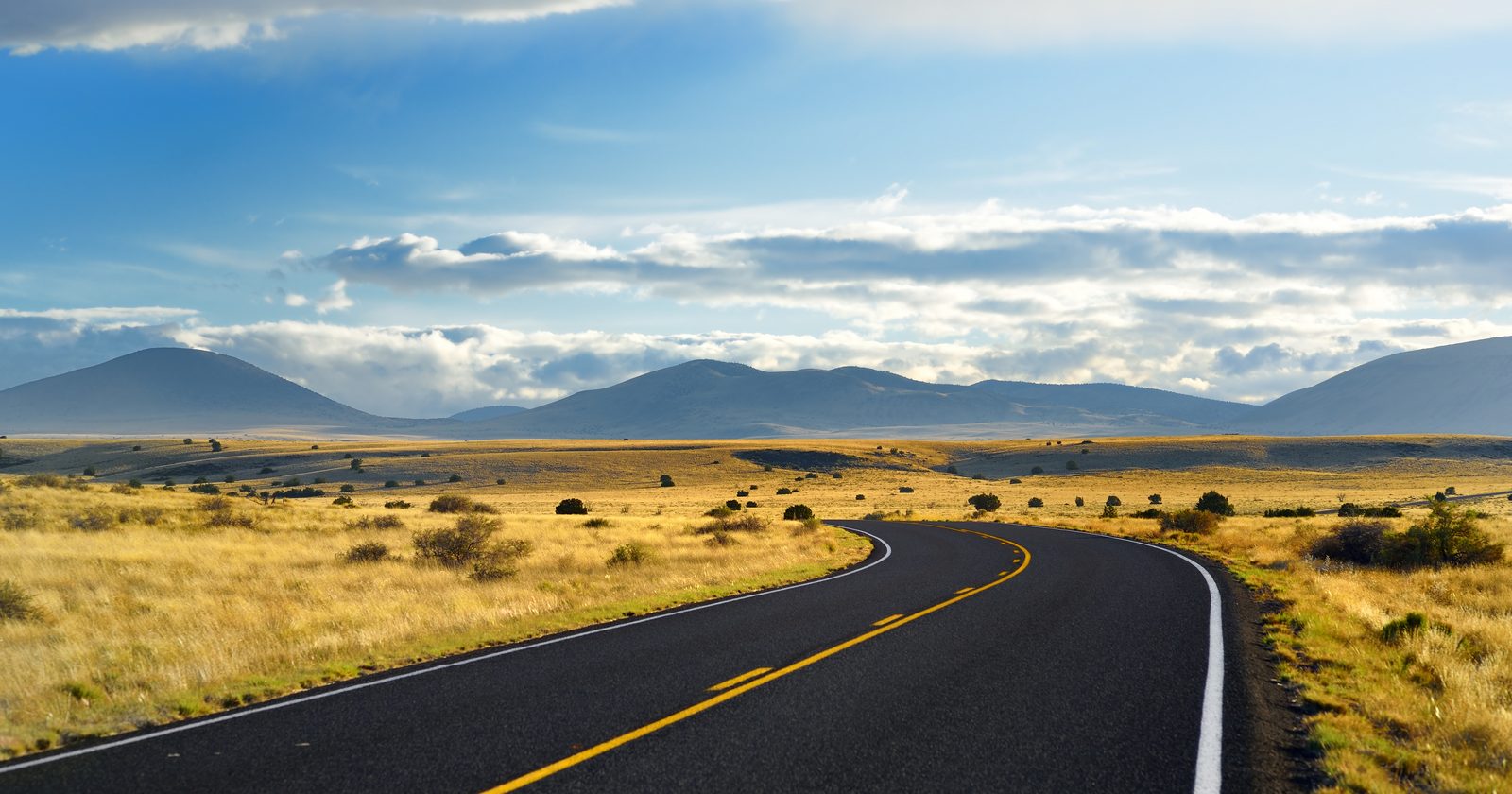 Open desert highway curving through golden high plains toward distant mountains